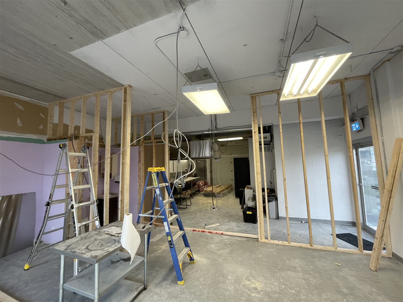 Commercial interior during the work, with exposed wood stud framing, ladders and work lights, and a range hood visible in the back room