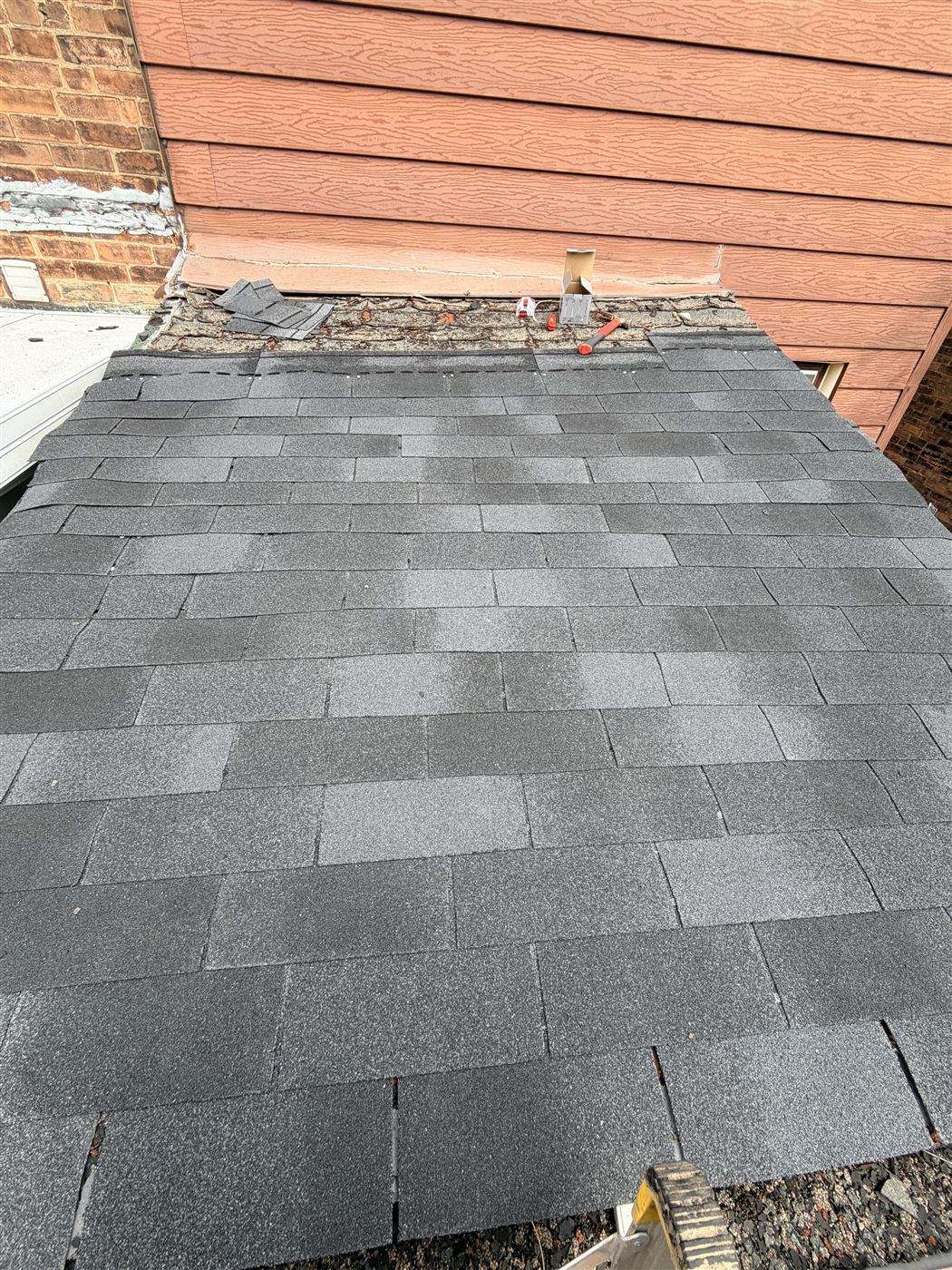 Lean-to roof with new grey asphalt shingles being installed against the siding of a brick building